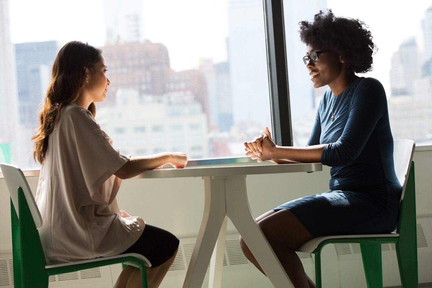 Two filmmakers in conversation at a table by a city window